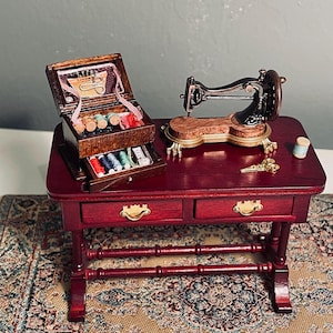 Könnte beinhalten: Ein Miniatur-Nähtisch aus dunklem Holz mit zwei Schubladen, ausgestattet mit einer schwarzen Nähmaschine aus Metall im Vintage-Stil, einer Nähkiste aus Holz mit offenem Deckel und Schublade voller bunter Garnrollen, einer kleinen goldenen Schere.