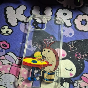 Puede incluir: Una funda de teléfono transparente con una ilustración pintada a mano de una cucaracha marrón sentada en un barril de madera en una mesa roja, sosteniendo una pequeña hamburguesa amarilla y verde en el centro de la cubierta.
