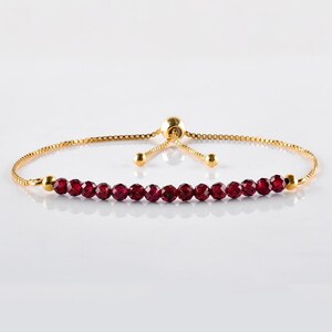 May include: A gold-colored adjustable bracelet featuring a row of faceted, deep red gemstones. The bracelet has a delicate chain and a sliding bead closure for a customizable fit. The gemstones are evenly spaced along the bracelet's center.