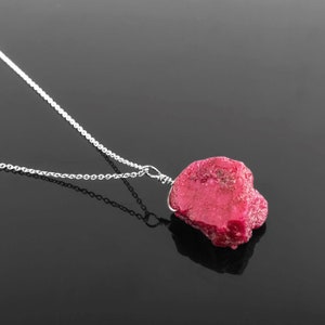 May include: A silver chain necklace with a raw, pink ruby pendant. The ruby is a rough, irregular shape and is set in a silver wire.