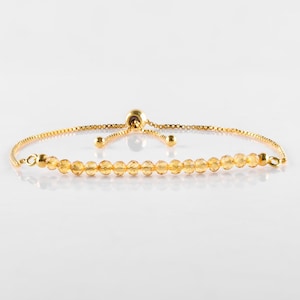 May include: A gold-colored bracelet with a delicate chain and a row of small, faceted, yellow-orange gemstones. The bracelet has an adjustable sliding clasp and is set against a white background.