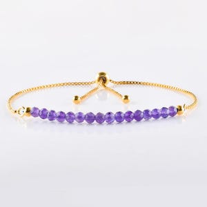 May include: A gold-toned bracelet featuring a row of small, faceted purple beads. The bracelet has a delicate chain and a sliding bead closure for adjustable sizing. The beads are evenly spaced, creating a simple yet elegant design.