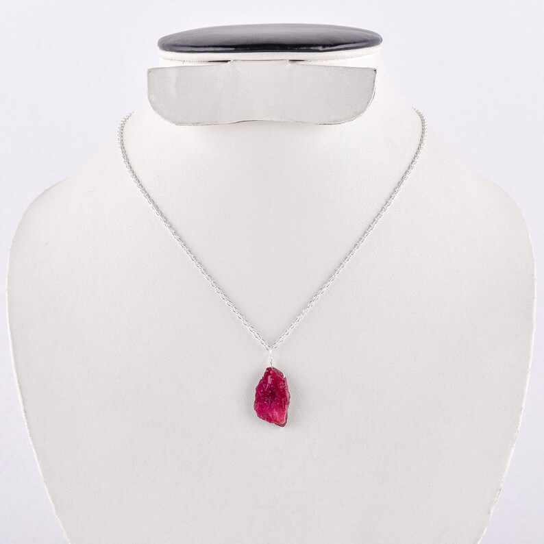 May include: A silver chain necklace with a raw, red ruby pendant. The ruby is a deep red color and has a natural, unpolished look.