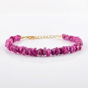 May include: A magenta-colored bracelet featuring raw, uncut ruby stones. The stones are strung together with a gold-colored chain and clasp. The bracelet is displayed on a white background.