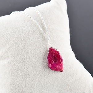 Könnte beinhalten: Eine Halskette aus Silber mit einem rohen, rosafarbenen Rubin-Anhänger. Der Rubin ist ein natürlicher, grob geschliffener Stein mit einer tiefen, satten Farbe.