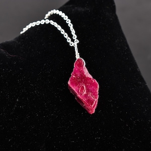 May include: A silver chain necklace with a raw ruby pendant. The ruby is a deep red color and has a rough, natural texture.