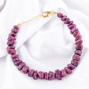 May include: A pink ruby gemstone bracelet with a gold clasp. The bracelet is made of small, irregular shaped stones.