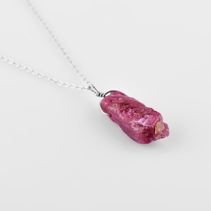 May include: A silver chain necklace featuring a raw, pink ruby pendant. The ruby is a natural, rough-cut stone with a deep pink hue.