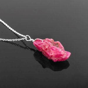 May include: A silver chain necklace with a raw pink ruby pendant. The ruby is a rough, natural stone with a deep pink colour.