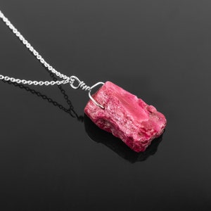 Könnte beinhalten: Eine silberne Kettenkette mit einem rosafarbenen, rohen Rubin-Anhänger. Der Rubin ist mit einem silbernen Draht umwickelt an der Kette befestigt.