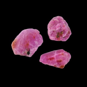 May include: Three pink ruby gemstones, each with a unique shape and texture. The gemstones are isolated against a black background.