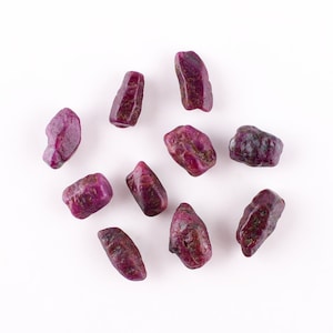 May include: A collection of ten small, rough, pink ruby gemstones on a white background.