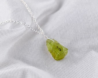 May include: A silver chain necklace with a raw, green peridot pendant. The peridot is a natural, rough stone with a light green color.