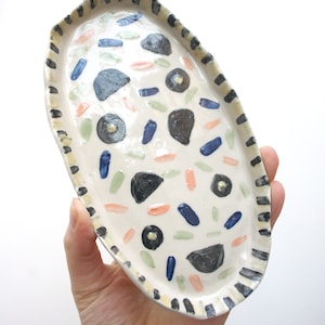 May include: White oval ceramic dish with a black and white striped rim. The dish is decorated with hand-painted blue, green, and pink abstract shapes.