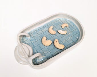 Puede incluir: Una bandeja de cerámica con forma de piscina, interior de azulejos azul claro y borde blanco. La bandeja presenta una escalera blanca esculpida en un lado y contiene cuatro anacardos de color beige claro sobre el fondo de la piscina.