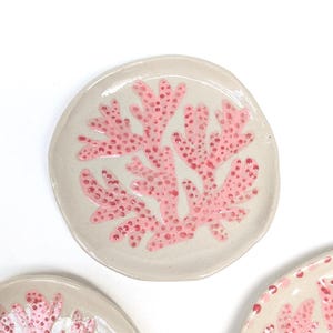 Könnte beinhalten: Eine runde, cremeweiße Keramikschale mit einem handgemalten rosa Korallenmotiv und strukturierten, erhabenen roten Punkten. Die Oberfläche hat ein glänzendes Finish und unregelmäßige, organische Ränder, die eine gesprenkelte Tonstruktur aufweisen.