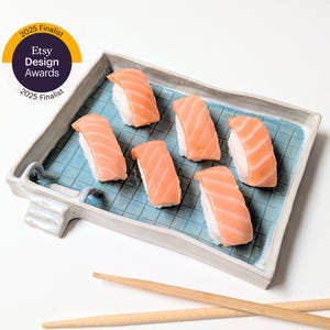 Peut inclure: Six pièces de sushi au saumon disposées sur une assiette rectangulaire en céramique bleu clair avec un motif quadrillé. Le sushi est garni de saumon orange vif. Deux baguettes en bois sont au premier plan. L'image comprend un badge "2025 Finalist Etsy Design Awards".