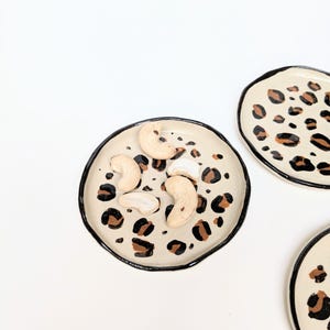 May include: A set of three small ceramic dishes with a leopard print design. The dishes are white with black outlines and brown spots. One dish has cashew nuts in it.