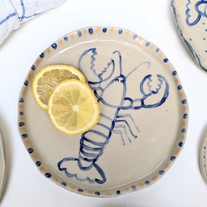 Puede incluir: Un plato de cerámica con un diseño de langosta azul pintado a mano. El plato tiene una base de color crema con un borde festoneado decorado con puntos azules y melocotón. Dos rodajas de limón descansan en el plato. Se ven platos adicionales en el fondo.