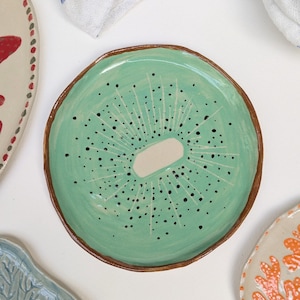 May include: A handmade ceramic plate with a kiwi fruit design. The plate features a light green center with black speckles and a white oval, mimicking the fruit's core. The rim is a dark brown. Other plates and a blue and white checkered cloth are in the background.