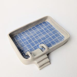 Puede incluir: Una bandeja de cer&aacute;mica rectangular con forma de piscina, con borde beige claro y un patr&oacute;n de cuadr&iacute;cula azul y blanco en la base interior. La bandeja presenta una peque&ntilde;a escalera de tres pelda&ntilde;os en un lado con un pasamanos curvo.