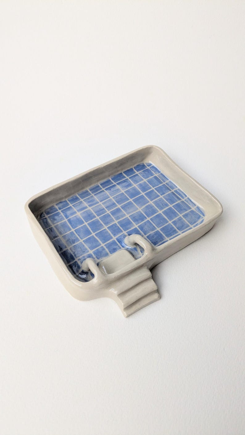 Puede incluir: Una bandeja de cer&aacute;mica rectangular con forma de piscina, con borde beige claro y un patr&oacute;n de cuadr&iacute;cula azul y blanco en la base interior. La bandeja presenta una peque&ntilde;a escalera de tres pelda&ntilde;os en un lado con un pasamanos curvo.