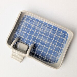 Puede incluir: Una jabonera rectangular de cer&aacute;mica con forma de piscina, con borde gris claro e interior azul con un patr&oacute;n de cuadr&iacute;cula blanco. La pieza incluye un peque&ntilde;o detalle de escalera adosado en un lado, imitando un dise&ntilde;o de piscina.