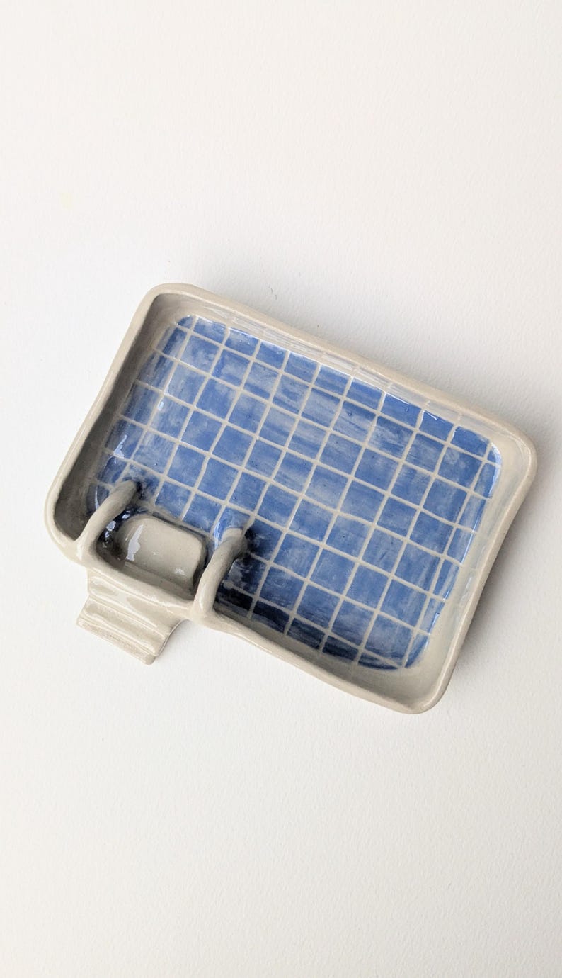 Puede incluir: Una jabonera rectangular de cer&aacute;mica con forma de piscina, con borde gris claro e interior azul con un patr&oacute;n de cuadr&iacute;cula blanco. La pieza incluye un peque&ntilde;o detalle de escalera adosado en un lado, imitando un dise&ntilde;o de piscina.
