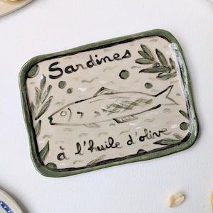 Op de afbeelding: Een rechthoekig keramieken dienblad met een groene rand en een beige middenstuk. Het dienblad is voorzien van een handgeschilderde illustratie van een vis, olijftakken en de tekst "Sardines à l'huile d'olive" in zwarte letters.