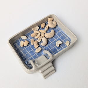 Puede incluir: Una bandeja rectangular de cer&aacute;mica con borde gris claro y un interior azul con un patr&oacute;n de cuadr&iacute;cula blanca. La bandeja tiene un peque&ntilde;o mango o soporte que sobresale por un lado y contiene una mezcla de cacahuetes y anacardos tostados.