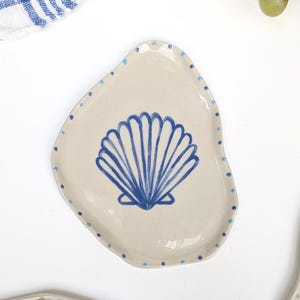 Può includere: Un piattino in ceramica beige dalla forma organica irregolare. Il centro presenta un disegno di una conchiglia di capasanta dipinto a mano in blu. Il bordo è decorato con una serie di piccoli punti blu chiaro e scuro lungo tutto il perimetro.