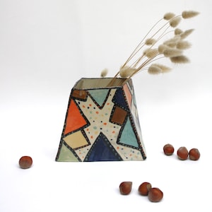Può includere: Un vaso in ceramica geometrica con un colorato design patchwork, contenente erba ornamentale essiccata. Il vaso presenta forme arancioni, blu, marroni e verdi con dettagli a pois. Diverse nocciole sono sparse.