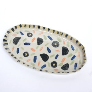 May include: An oval ceramic serving tray with a white background and a black, yellow, blue, green, and orange abstract pattern.