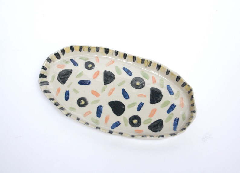 May include: An oval ceramic serving tray with a white background and a black, yellow, blue, green, and orange abstract pattern.