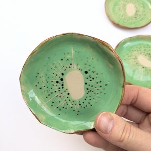 Puede incluir: Platos de cerámica hechos a mano con forma de rodajas de kiwi. Los platos presentan un exterior verde claro con un borde marrón y un centro beige, imitando el diseño de la fruta. Pequeños puntos negros están dispersos por la superficie verde.