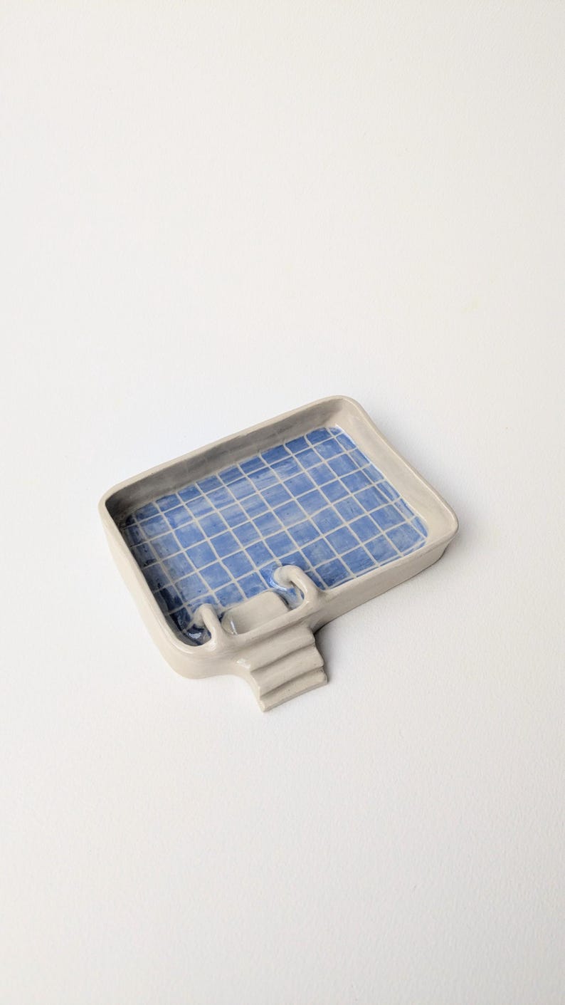 Puede incluir: Una bandeja rectangular de cer&aacute;mica con exterior gris claro y un patr&oacute;n de cuadr&iacute;cula azul y blanco en la base interior, dise&ntilde;ada para parecerse a una piscina. La bandeja cuenta con una peque&ntilde;a escalera adjunta en un lado.