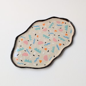 Può includere: Un vassoio in ceramica dalla forma organica irregolare con una base color crema. La superficie presenta un motivo sparso di piccoli semicerchi rosa, forme frastagliate azzurre, triangoli arancioni, linee blu ondulate e brevi tratti bianchi e neri.