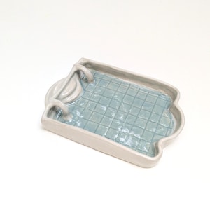 Può includere: Un vassoio rettangolare in ceramica a forma di piscina con bordo bianco lucido e interno azzurro caratterizzato da un motivo a griglia. Il vassoio include una piccola scala scolpita su un lato, che imita l'aspetto di una vasca.