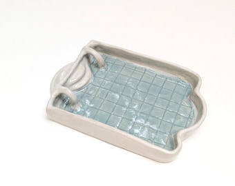 Puede incluir: Una bandeja de cerámica rectangular con forma de piscina, con un borde blanco brillante y un interior azul claro con un patrón de cuadrícula. La bandeja incluye una pequeña escalera esculpida en un lado, imitando un estanque.