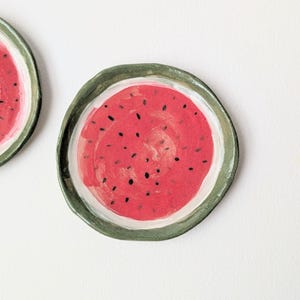 Op de afbeelding: Twee ronde keramische schaaltjes met onregelmatige, met de hand gevormde randen. Het midden van elk schaaltje heeft een felroze watermeloenontwerp met verspreide zwarte pitjes, omringd door een dikke, donkergroene rand op een witte achtergrond.