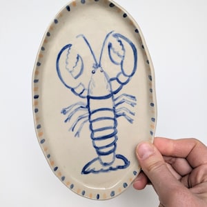 Puede incluir: Un plato ovalado de cerámica blanca con un diseño de langosta pintado a mano en azul y blanco. El plato tiene un borde de puntos naranja claro y azules.