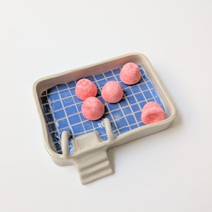 Puede incluir: Una fuente de cerámica con un patrón de baldosas azules y un pequeño detalle de escalera. La fuente contiene cinco caramelos rosas y redondos. La fuente tiene un borde gris y está sobre un fondo blanco.