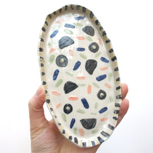 May include: A white oval ceramic dish with a black and yellow striped border. The dish is decorated with hand-painted blue, green, pink, and black abstract shapes.