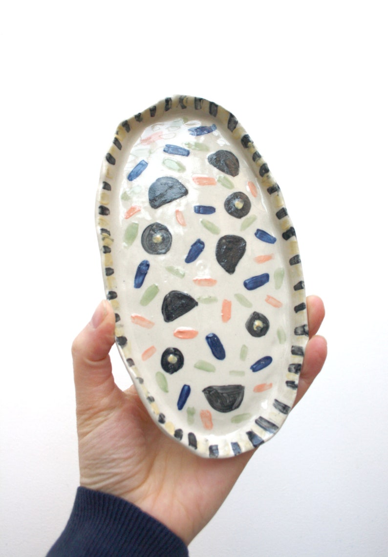 May include: A white oval ceramic dish with a black and yellow striped border. The dish is decorated with hand-painted blue, green, pink, and black abstract shapes.