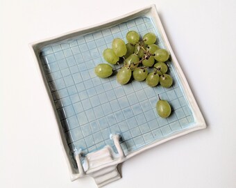 Puede incluir: Una fuente cuadrada de cerámica con un interior de baldosas azul claro, que se asemeja a una piscina. Un racimo de uvas verdes descansa sobre la fuente. La fuente tiene un borde blanco y una pequeña escalera de piscina decorativa blanca.