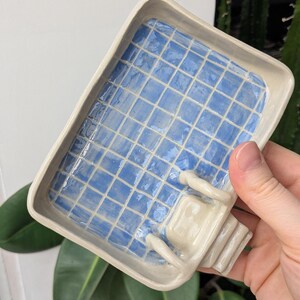 Puede incluir: Una bandeja rectangular de cer&aacute;mica con acabado brillante, que presenta un patr&oacute;n de cuadr&iacute;cula azul claro que imita el interior de una piscina. La bandeja incluye una peque&ntilde;a escalera blanca esculpida en tres dimensiones en un lado.