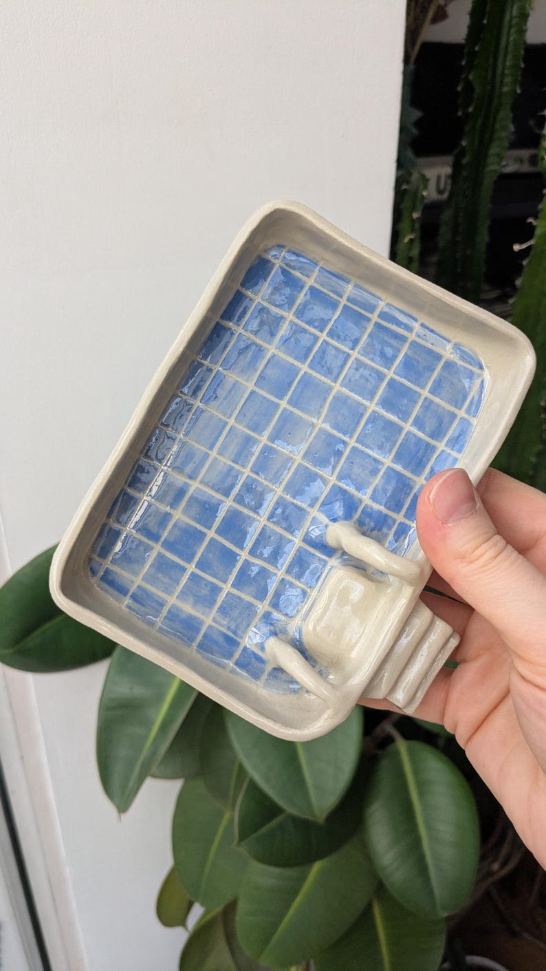 Puede incluir: Una bandeja rectangular de cer&aacute;mica con acabado brillante, que presenta un patr&oacute;n de cuadr&iacute;cula azul claro que imita el interior de una piscina. La bandeja incluye una peque&ntilde;a escalera blanca esculpida en tres dimensiones en un lado.