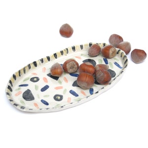 May include: A white ceramic oval serving tray with a colorful abstract pattern. The tray is filled with hazelnuts.