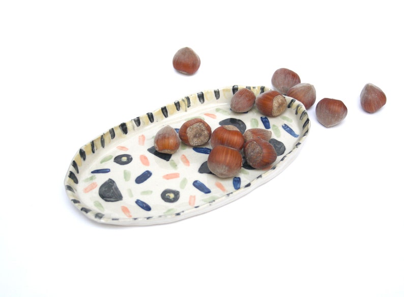 May include: A white ceramic oval serving tray with a colorful abstract pattern. The tray is filled with hazelnuts.