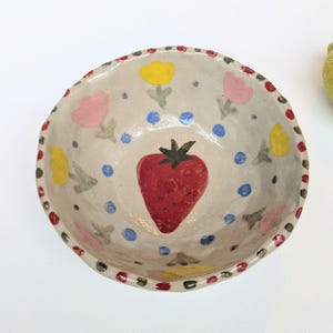May include: A small white ceramic bowl with a red strawberry, yellow and pink flowers, and blue dots painted on the inside. The bowl has a red and black scalloped edge.